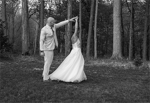 Groom twirls bride around in front of the woods.