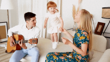 dad playing guitar with little girl jumping on the couch and mom holding her hand