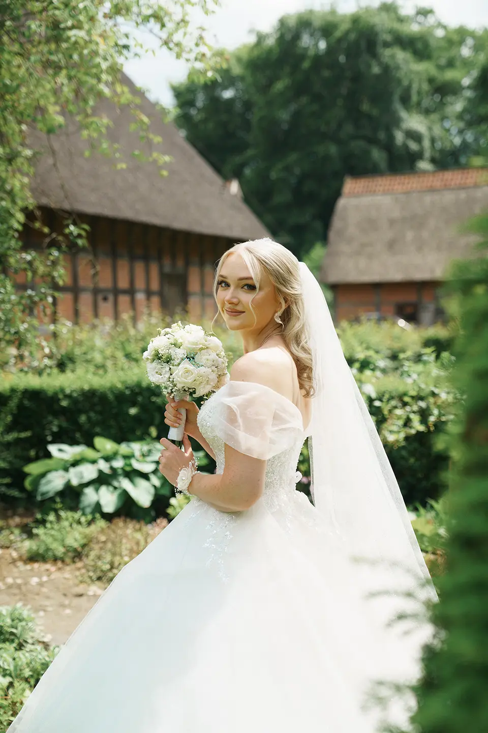 Braut im Off-Shoulder-Kleid hält weißen Strauß vor Fachwerk und Reetdächern; sanftes Licht betont natürliche hochzeitsfotografie, hochzeitsbilder draußen und hochzeitsfotografie mit stil