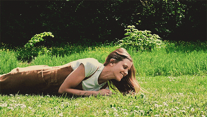 GIF of woman in jumpsuit rolling in grassy field and smiling at the camera
