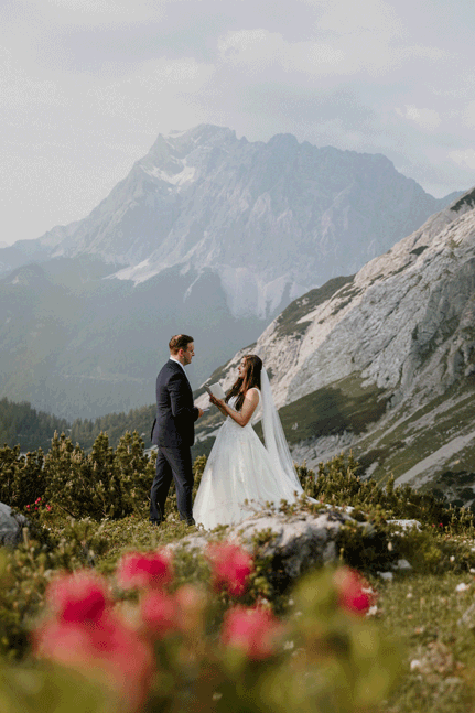 A lesbian couple in white wedding attire pop champagne. They are both smiling and laughing, the champagne is spraying and the mountain peaks of Triglav National Park in Slovenia are in the background.