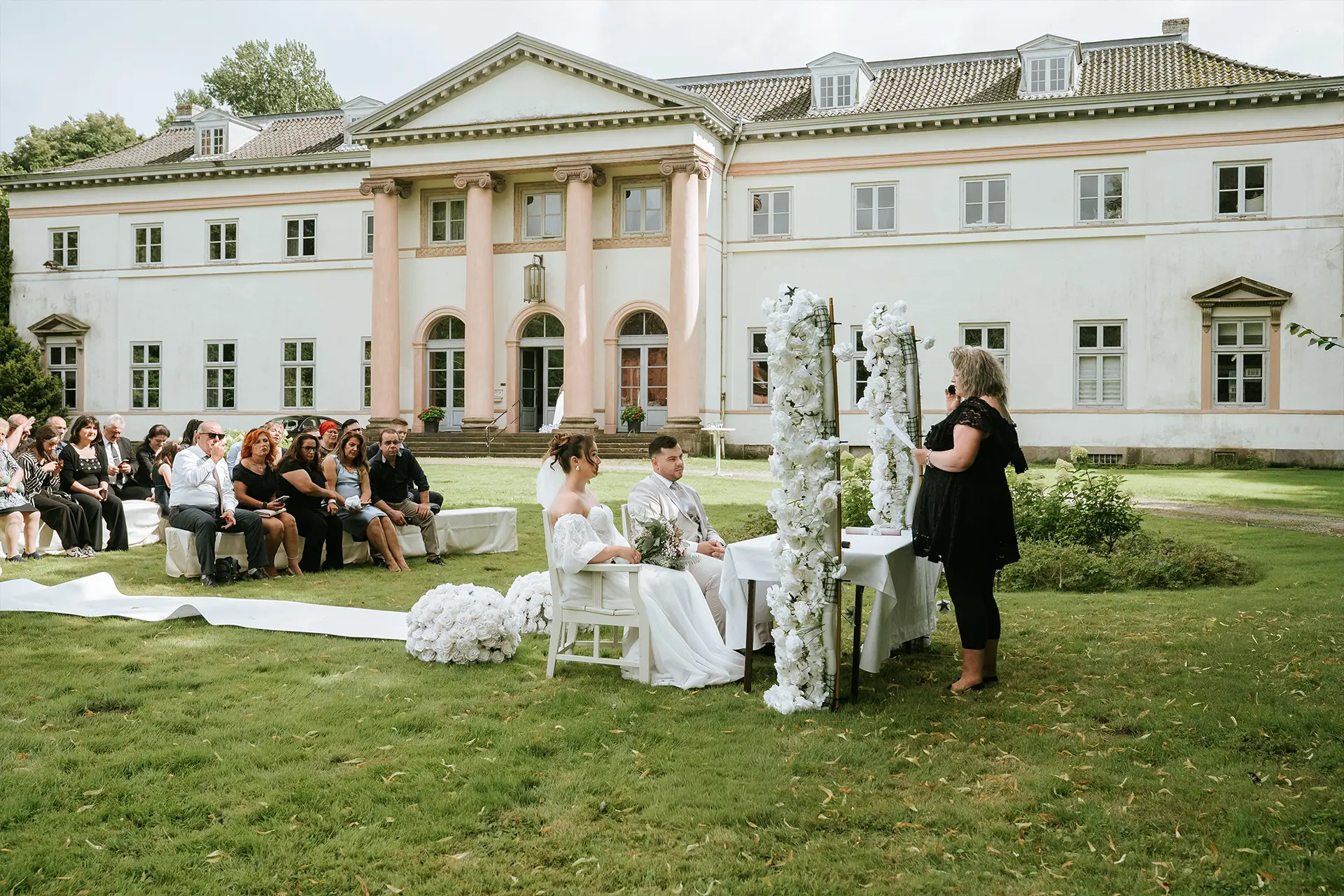 Brautpaar sitzt unter weißem Blütendekor, die Rednerin führt die Zeremonie; vor dem Herrenhaus entstehen natürliche hochzeitsfotografie, edle Hochzeitsreportage Kiel und feine reportagefotografie hochzeit