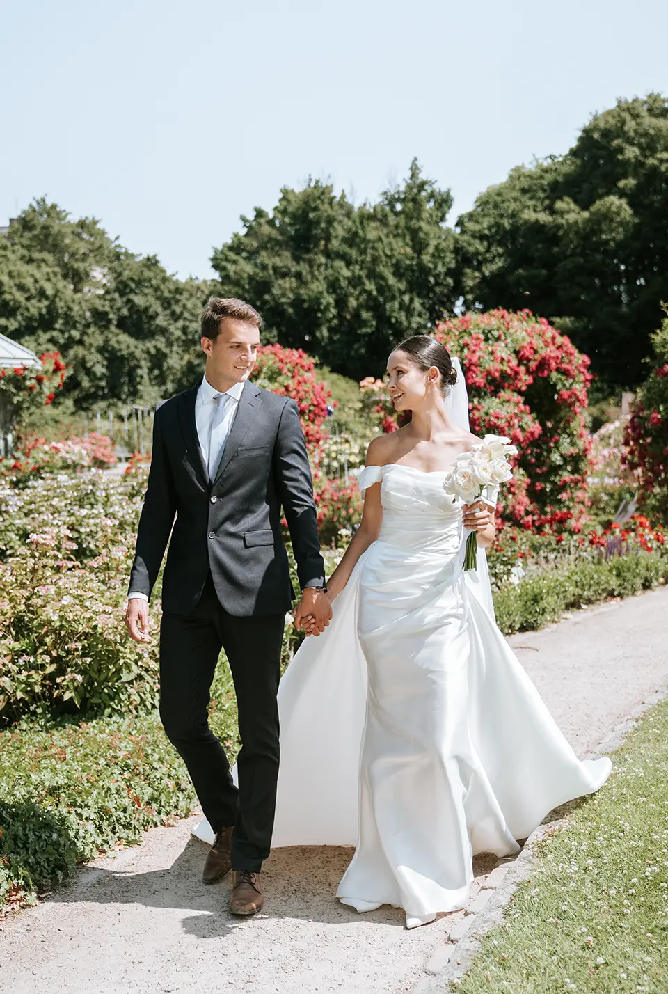 Braut und Bräutigam spazieren Hand in Hand durch den Rosengarten; Satinkleid und weißer Strauß unterstreichen natürliche hochzeitsfotografie und stimmungsvolle hochzeitsbilder draußen