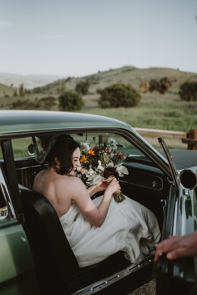 Bride sitting in the passenger seat of a vintage green car, holding her bouquet, as her husband opens the door