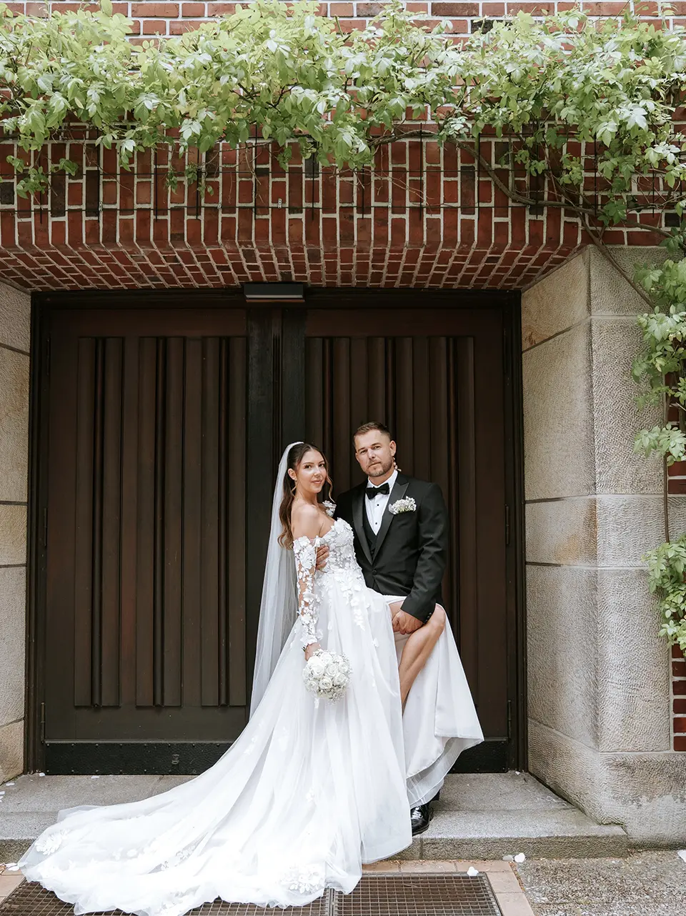 Braut und Bräutigam posieren vor dunklem Holztor unter Backsteinranken; die Braut mit langer Schleppe und Bouquet – natürliche Hochzeitsfotografie, Hochzeitsfotografie mit Stil in Hannover