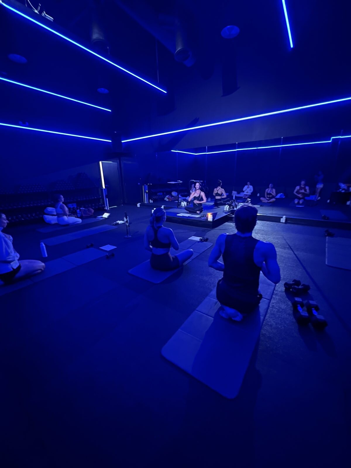 Breathwork and meditation practice in a darkened studio lit by deep blue LED strips, with students seated cross-legged on mats facing a lead facilitator.