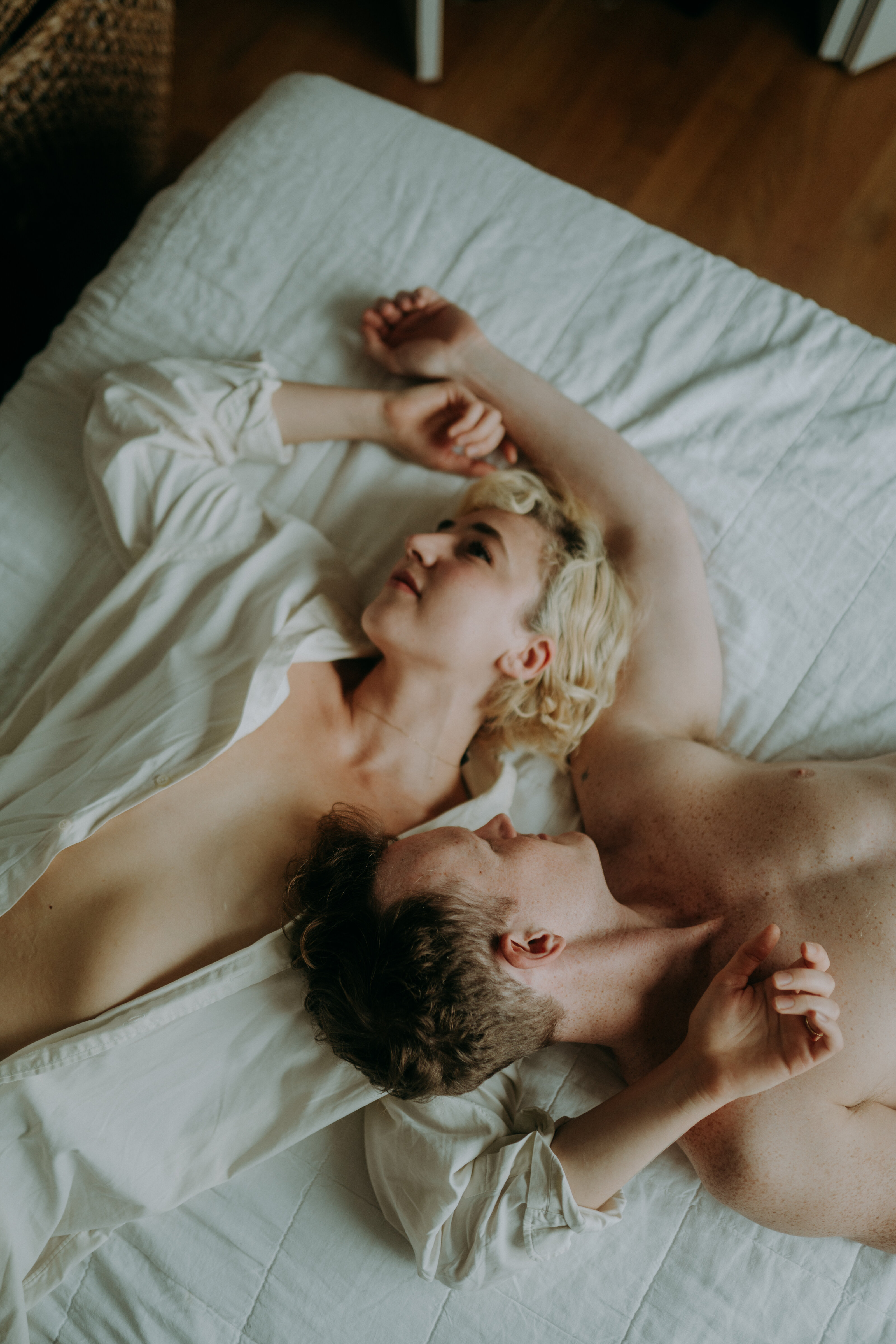A shirtless couple lies on a bed holding hands, their faces close and serene.