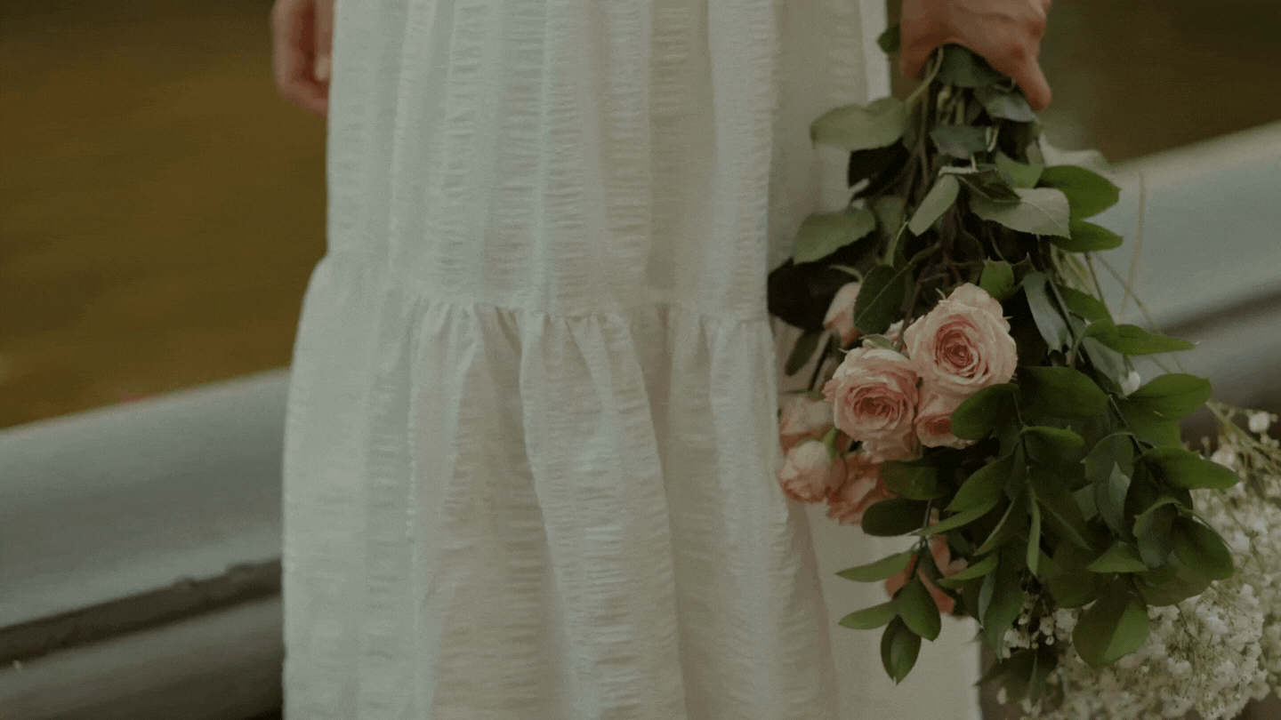 Moving image of woman in a white summery dress holding a bunch of blush pink roses and leafy plants