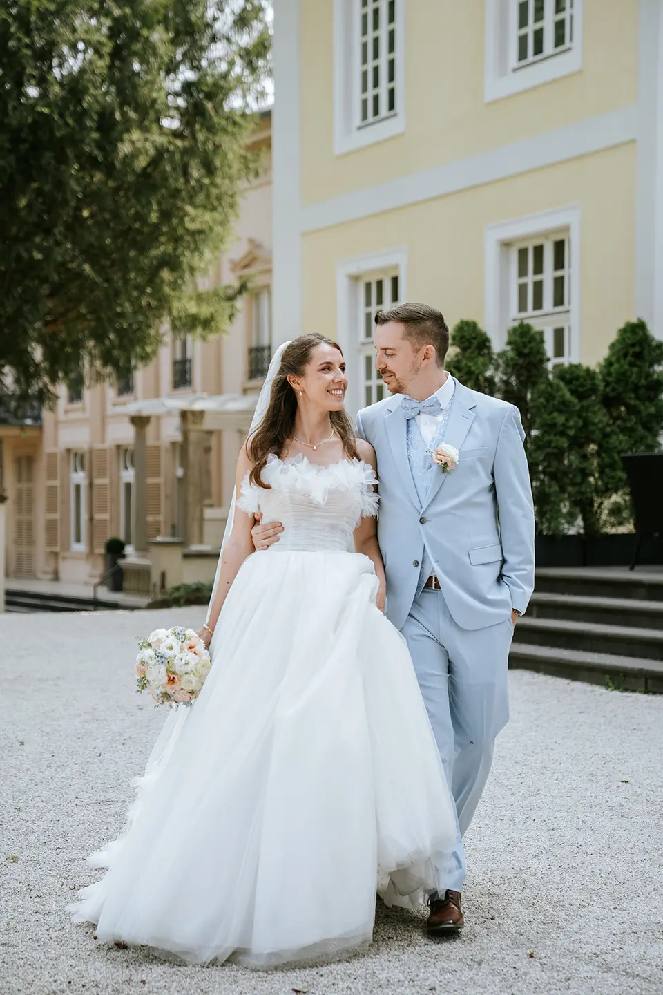 Brautpaar vor gelber Villa: Braut mit Bouquet und weitem Kleid, Bräutigam im hellblauen Anzug. Vertikales Mobil-Header-Bild – natürliche Hochzeitsfotografie, emotionale Hochzeitsbilder