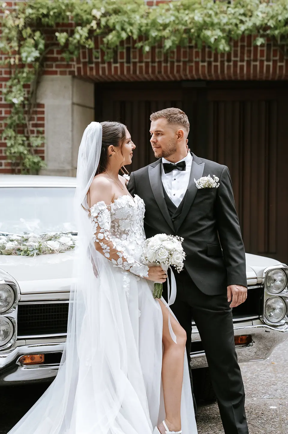Braut und Bräutigam stehen am geschmückten Oldtimer, halten den Strauß und sehen sich verliebt an; moderne hochzeitsfotografie und emotionale hochzeitsbilder