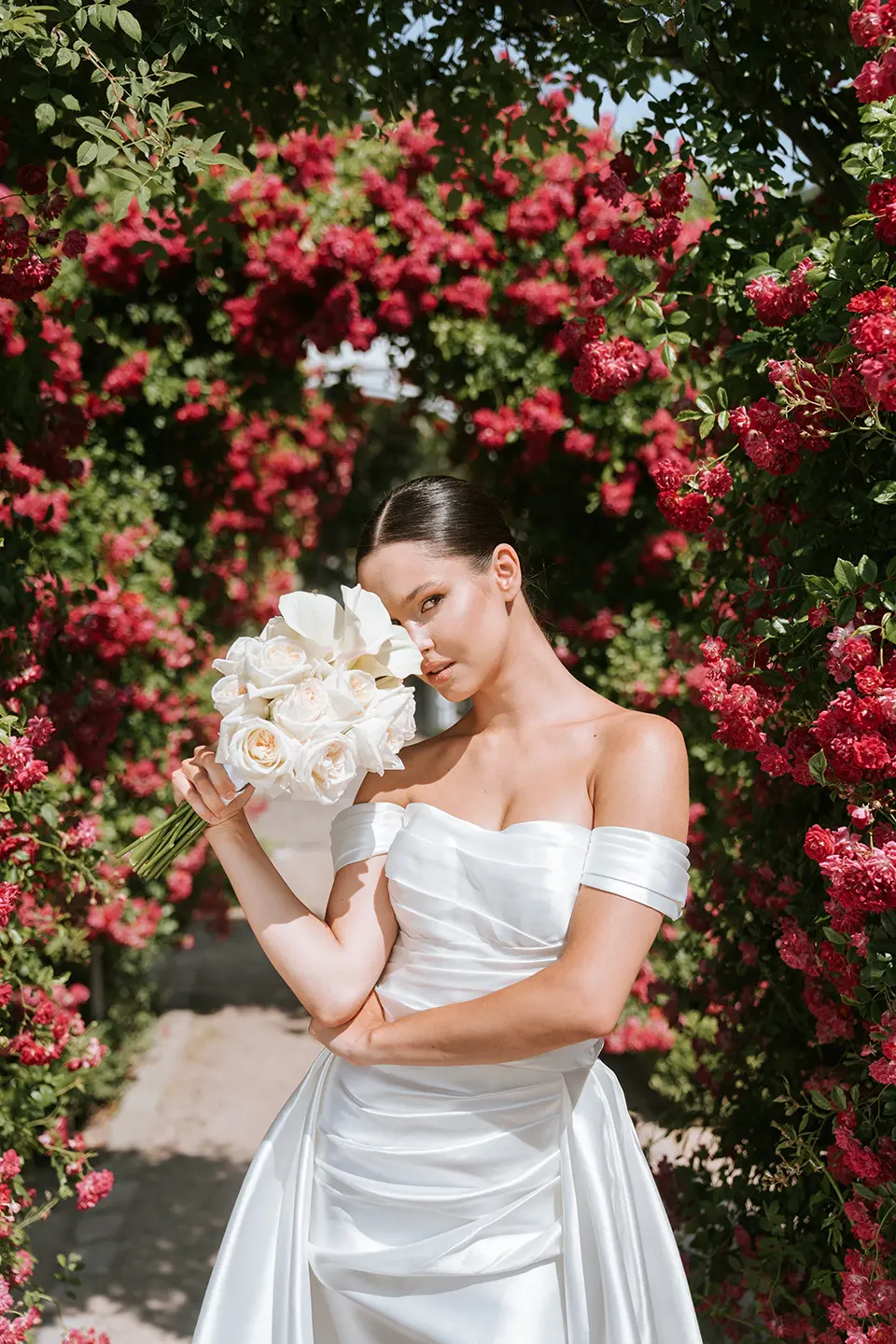 Vor leuchtenden Rosen hält die Braut ein weißes Bouquet und schaut leicht zur Seite. Künstlerische hochzeitsfotos treffen auf moderne hochzeitsfotografie, clean und elegant