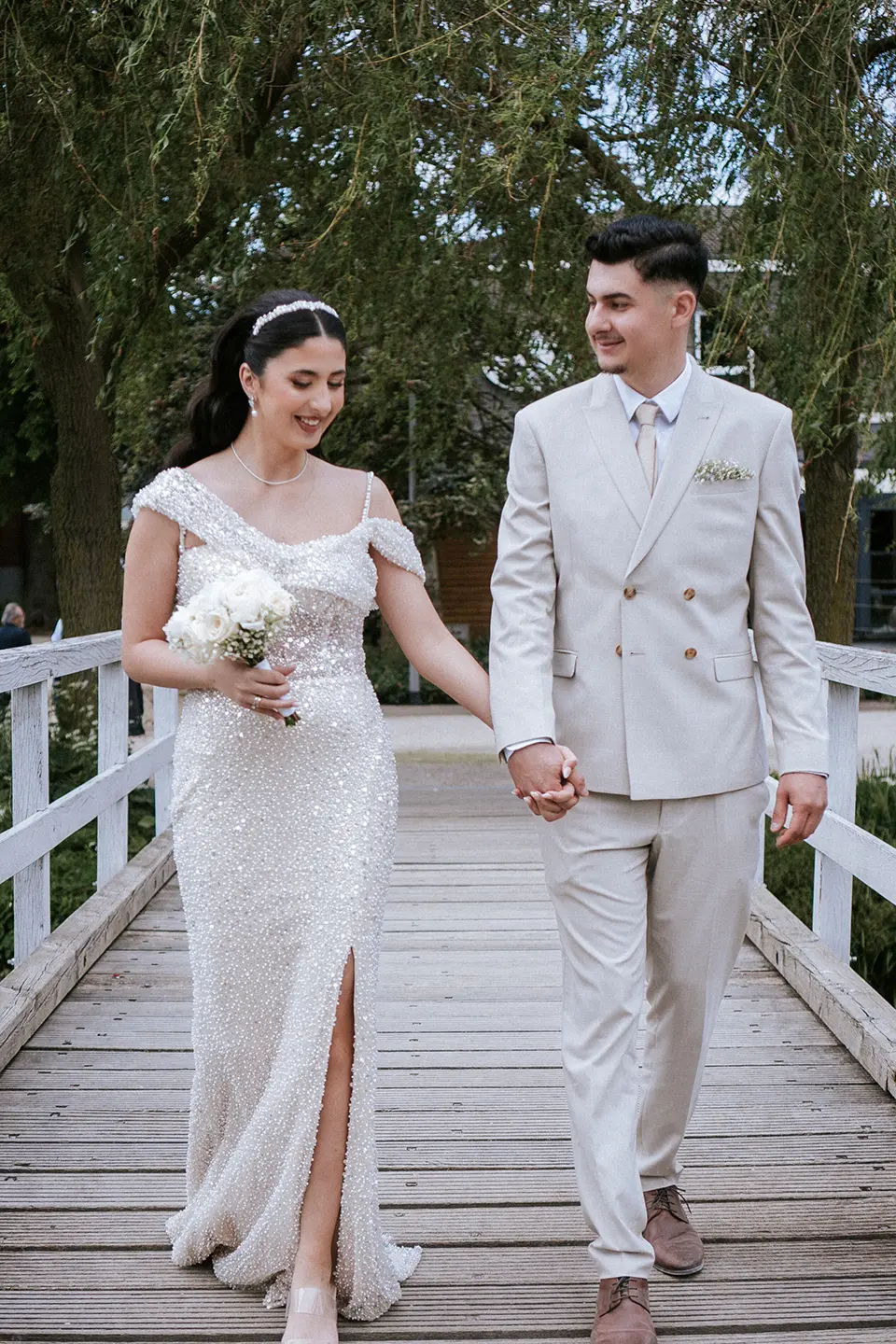 Braut in funkelndem Kleid und Bräutigam im hellen Anzug gehen Hand in Hand über eine hölzerne Parkbrücke; natürliche hochzeitsfotografie und hochzeitsbilder draußen