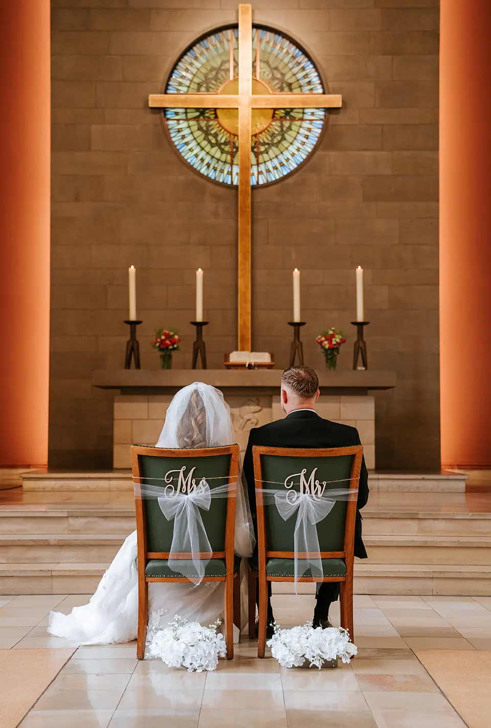 Vor dem Altar sitzen Braut und Bräutigam auf mit Schleifen gebundenen Stühlen, Kerzen und Kreuz bilden den Rahmen; ganztägige hochzeitsreportage und klassische hochzeitsfotos