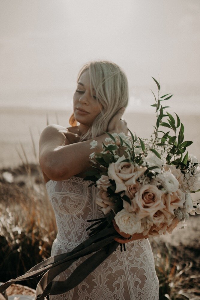 A bride in a strapless lace gown holds a large bouquet of blush roses and greenery on a sandy beach.