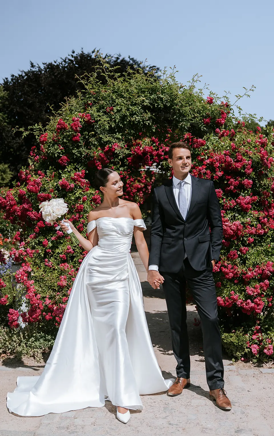 Vor einem üppigen Rosenbogen stehen Braut und Bräutigam Hand in Hand, die Braut hält den Strauß; klassische hochzeitsfotos in eleganter hochzeitsfotografie mit stil