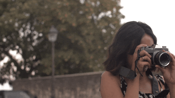 A photographer shoots pictures outdoors on her camera. She has shoulder length dark hair, and the camera pans back and forth as she moves around.