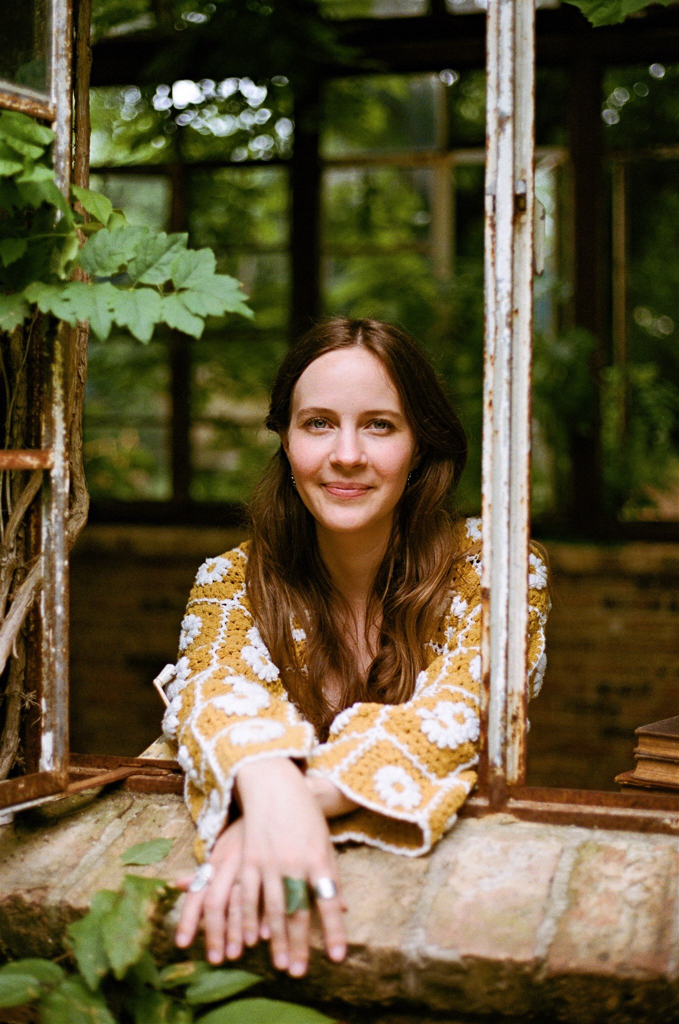 A woman with long brown hair, wearing a yellow floral-patterned top, leans on a brick windowsill in an old structure surrounded by greenery—perfect inspiration for brand and web design for small businesses.