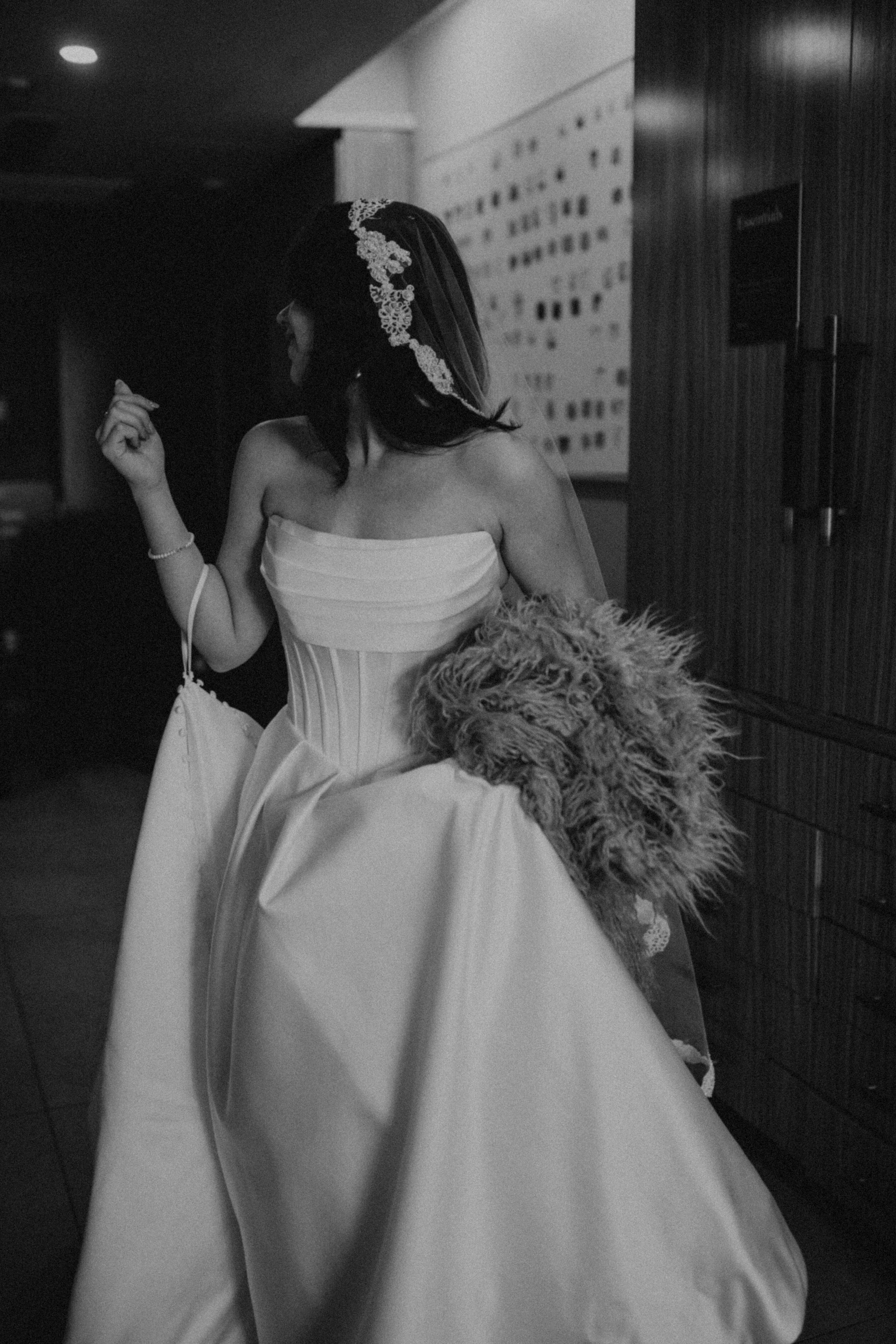 Bride in lace veil and strapless gown walks through a hallway holding her skirt and a textured wrap.