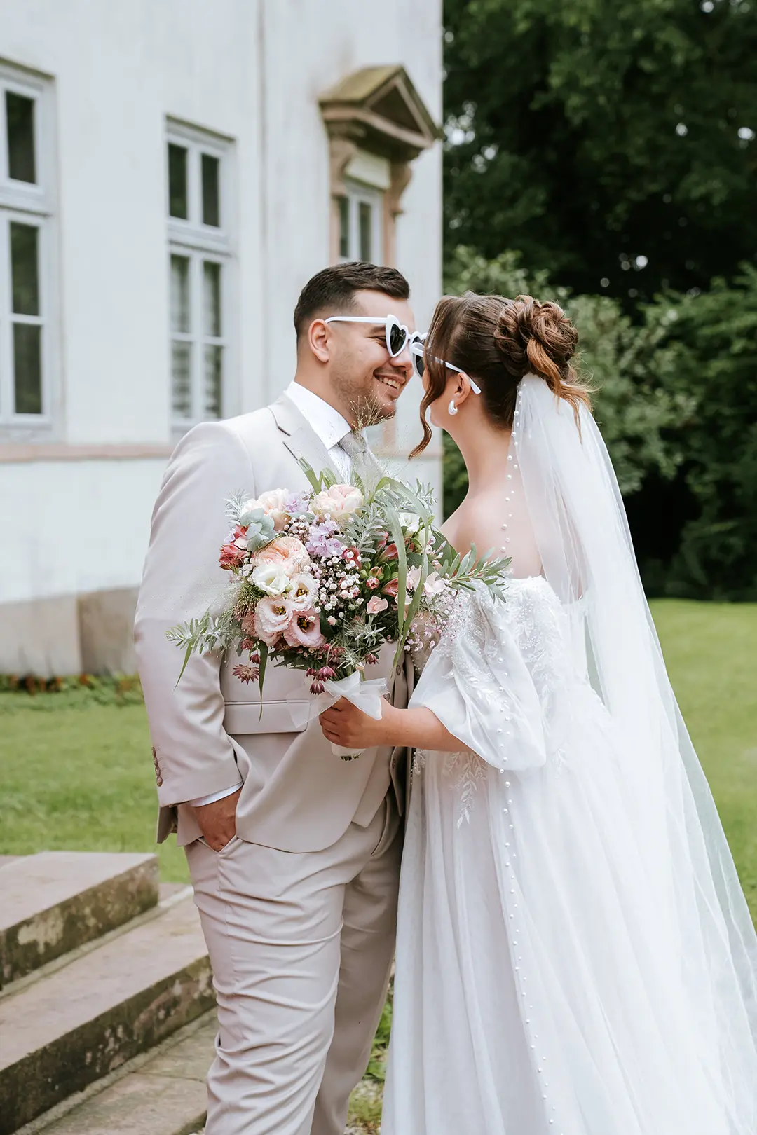 Das Paar lacht sich an, beide mit weißen Sonnenbrillen, die Braut hält ein üppiges Bouquet – moderne Hochzeitsfotografie trifft auf ein verspieltes Pärchenshooting mit emotionalen Hochzeitsbildern