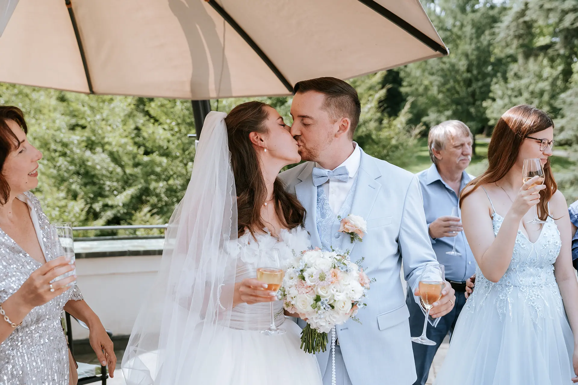 Beim Sektempfang unter dem Sonnenschirm küssen sich Braut und Bräutigam, Gäste stoßen mit Sekt an; moderne hochzeitsfotografie fängt emotionale hochzeitsbilder und klassische hochzeitsfotos ein.