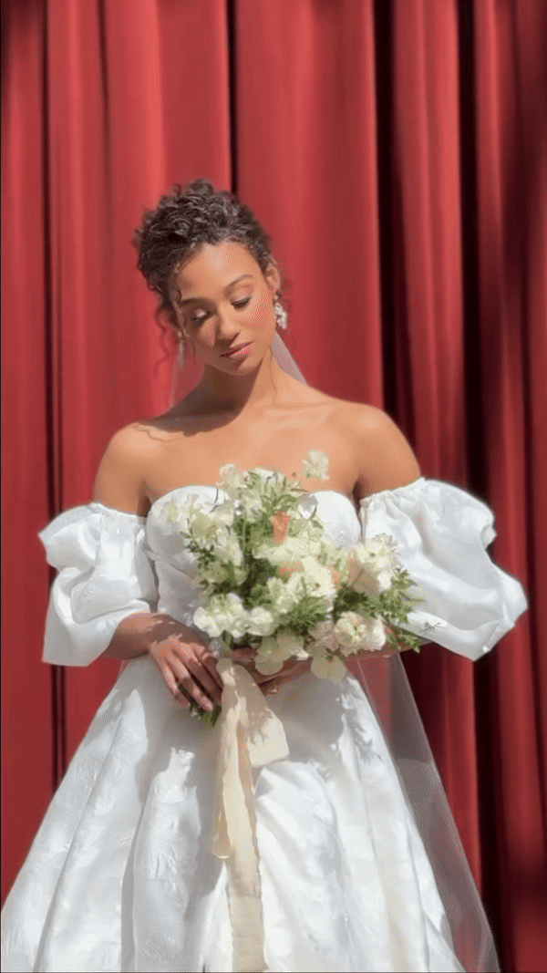 A bride in an elegant white dress holds a bouquet of white and green flowers, standing before rich red curtains, exuding a serene and joyful mood.