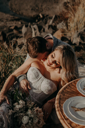 Groom kisses bride’s shoulder as she smiles, sitting together on a beach picnic setup with flowers.