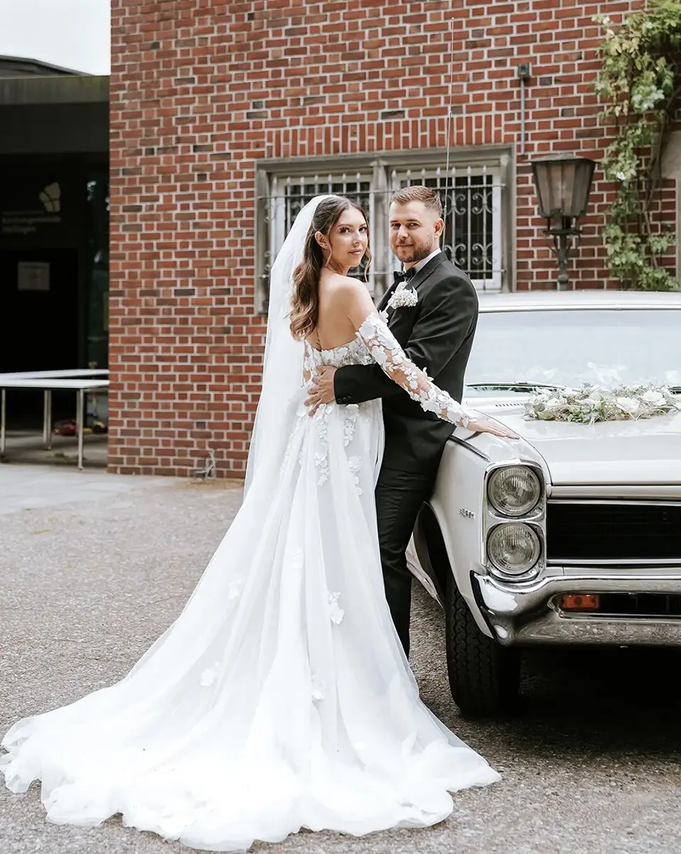 Die Braut zeigt ihre lange Schleppe am verzierten Oldtimer, während der Bräutigam sie sanft hält; klassische hochzeitsfotos treffen auf hochzeitsfotografie mit stil
