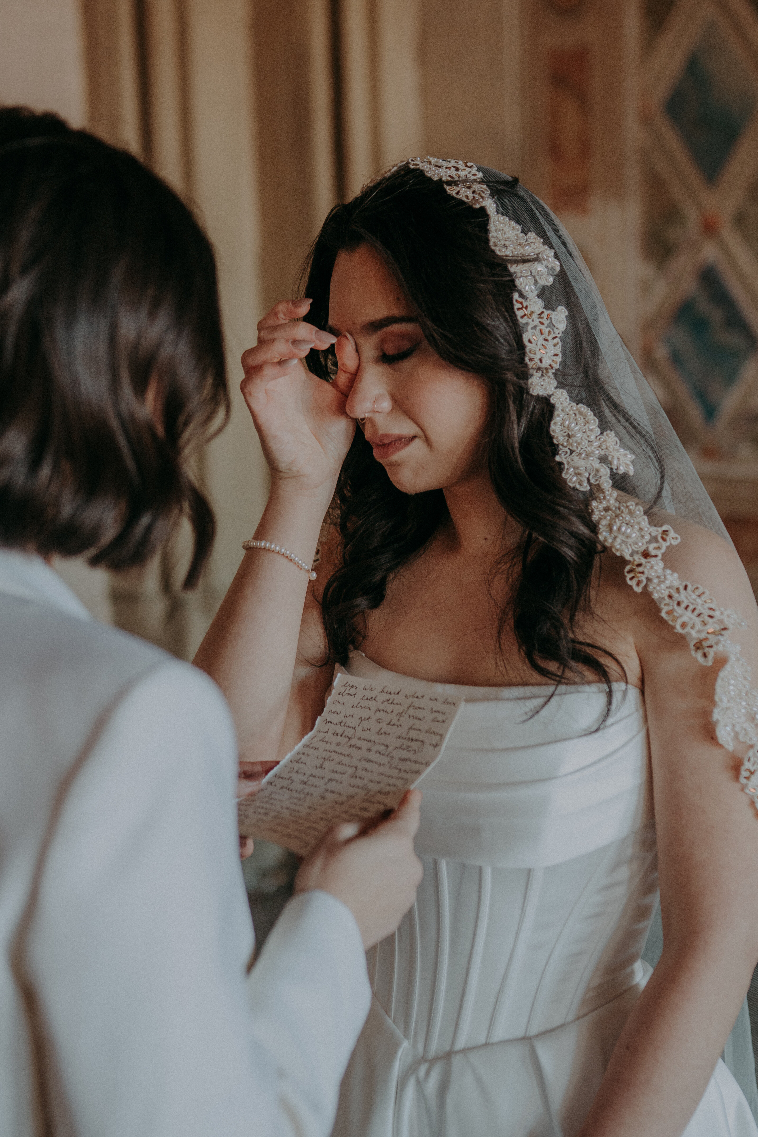 A bride wearing a veil wipes away a tear while another woman in a suit reads vows to her.