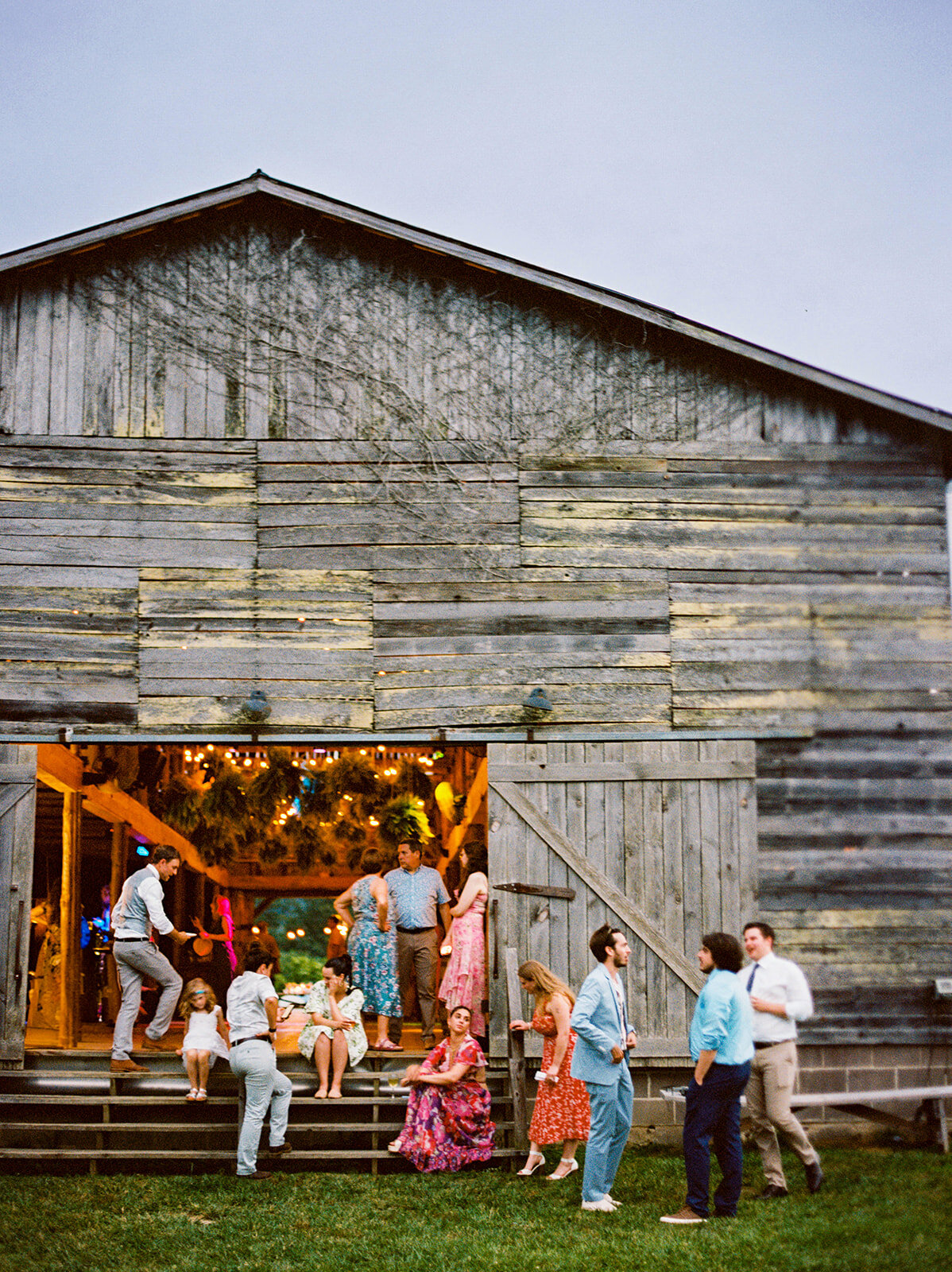 during the cocktail hour of a wedding, a number of guests sit outside a barn at Paint Rock Farm where the rest of the wedding is. Some guests are standing, some are seated, some are leaned against some stairs and look to be enjoying themselves