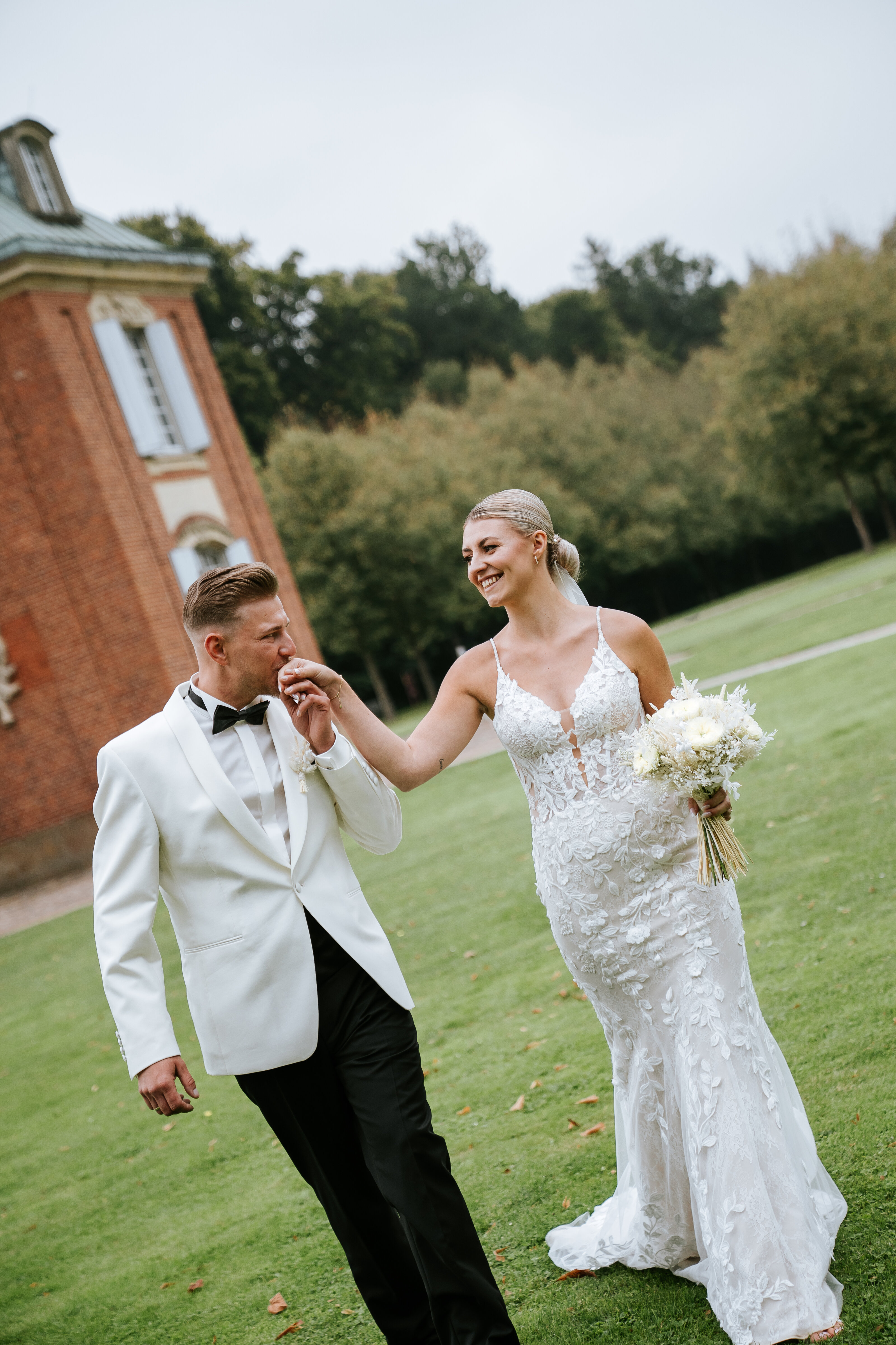 Bräutigam richtet der Braut zärtlich das Kleid, sie hält den Strauß zwischen hohen Hecken; warmer Gartenmoment für pärchenshooting hochzeit, natürliche hochzeitsfotografie und emotionale hochzeitsbilder