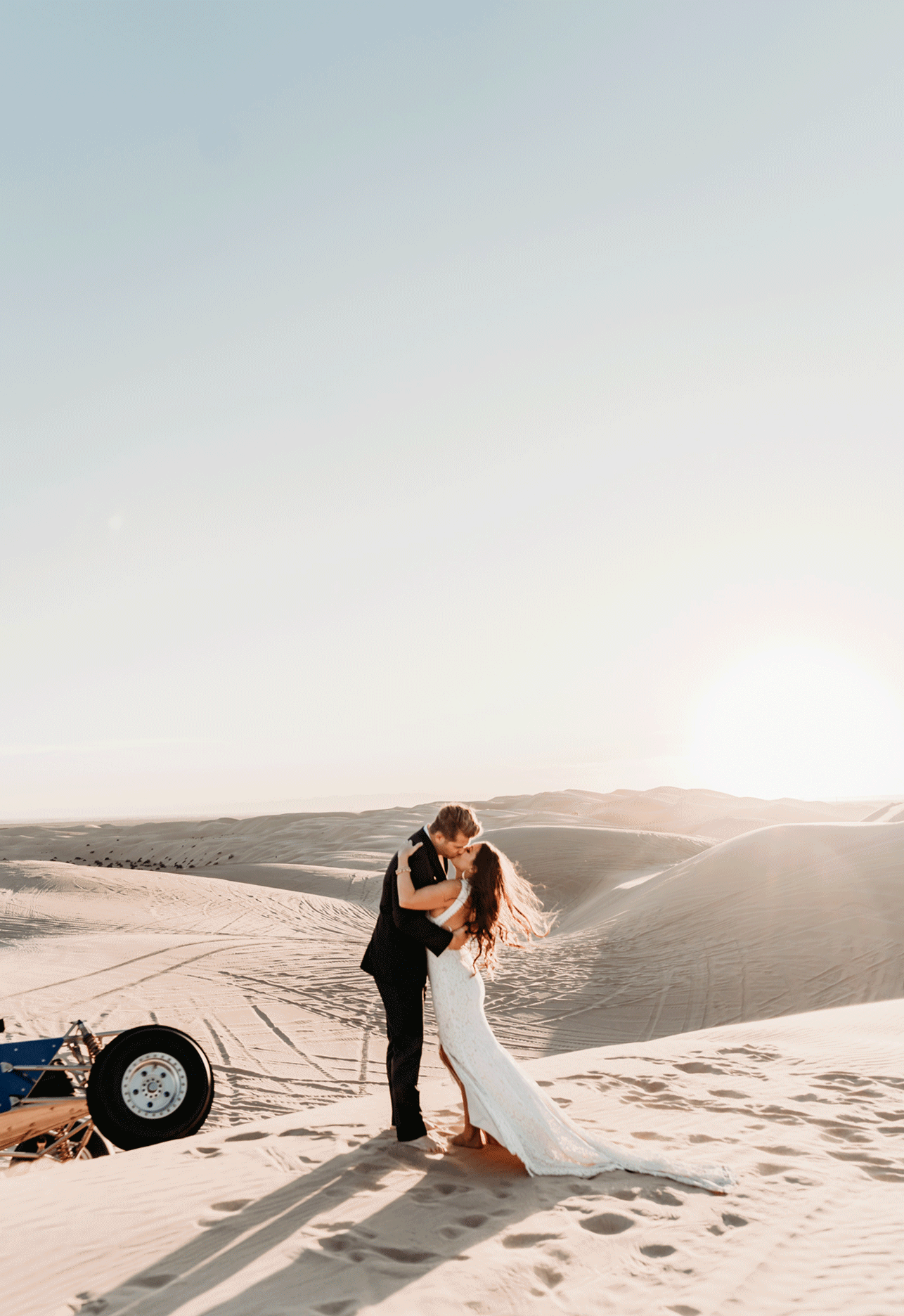 Bride and groom kiss as the sun goes down at Imperial Sand Dunes in Arizona after their wedding. A side by side flies behind them kicking up sand for a dramatic, smokey like effect