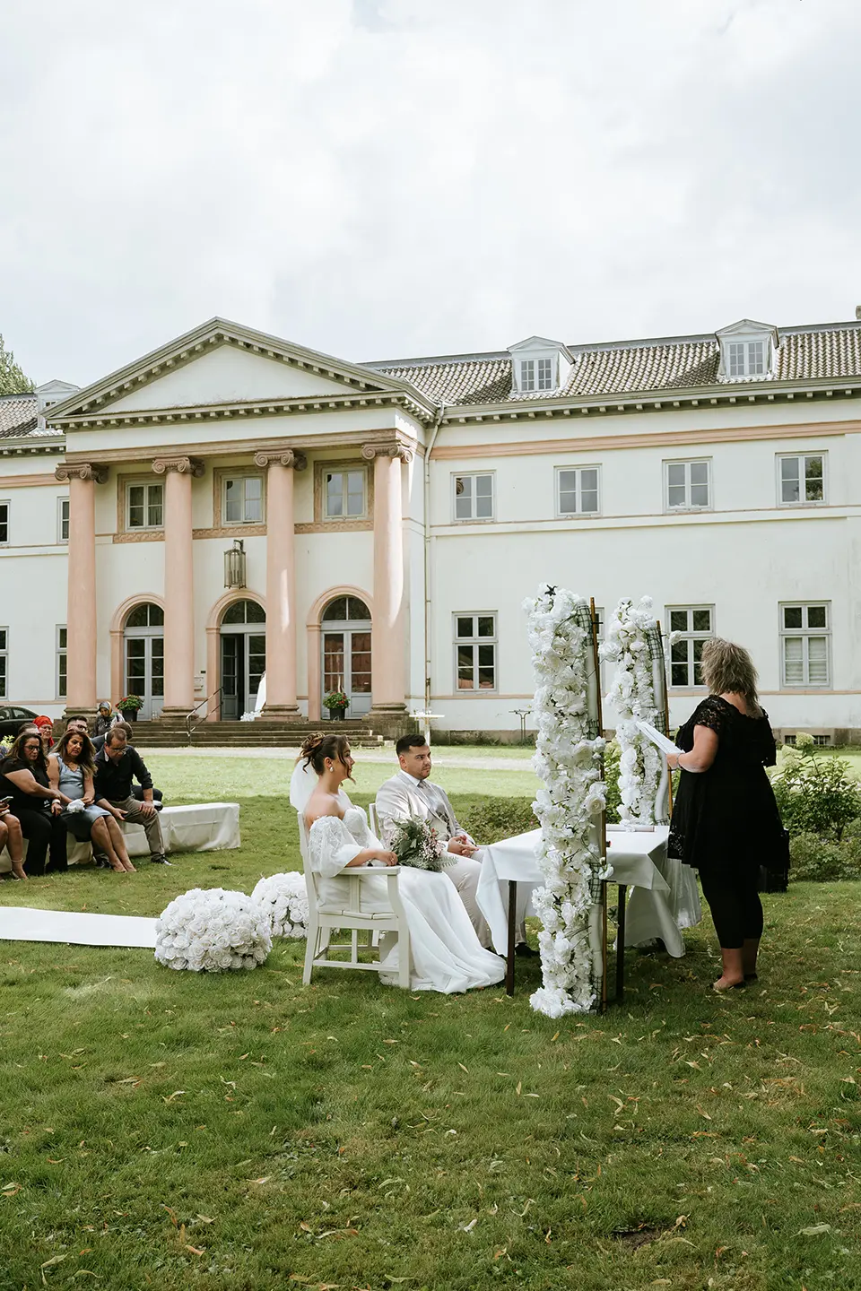 Freie Trauung vor klassischem Herrenhaus, das Paar sitzt der Rednerin gegenüber zwischen weißen Blumenbögen. Reportagefotografie hochzeit und hochzeitsfotograf für schloss-hochzeit begleiten die Szene