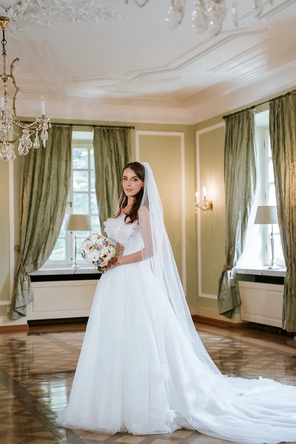 Stehendes Brautporträt im prunkvollen Saal mit Kronleuchtern, langer Schleppe und zartem Bouquet; klassische hochzeitsfotos in fine art hochzeitsfotografie Anmutung