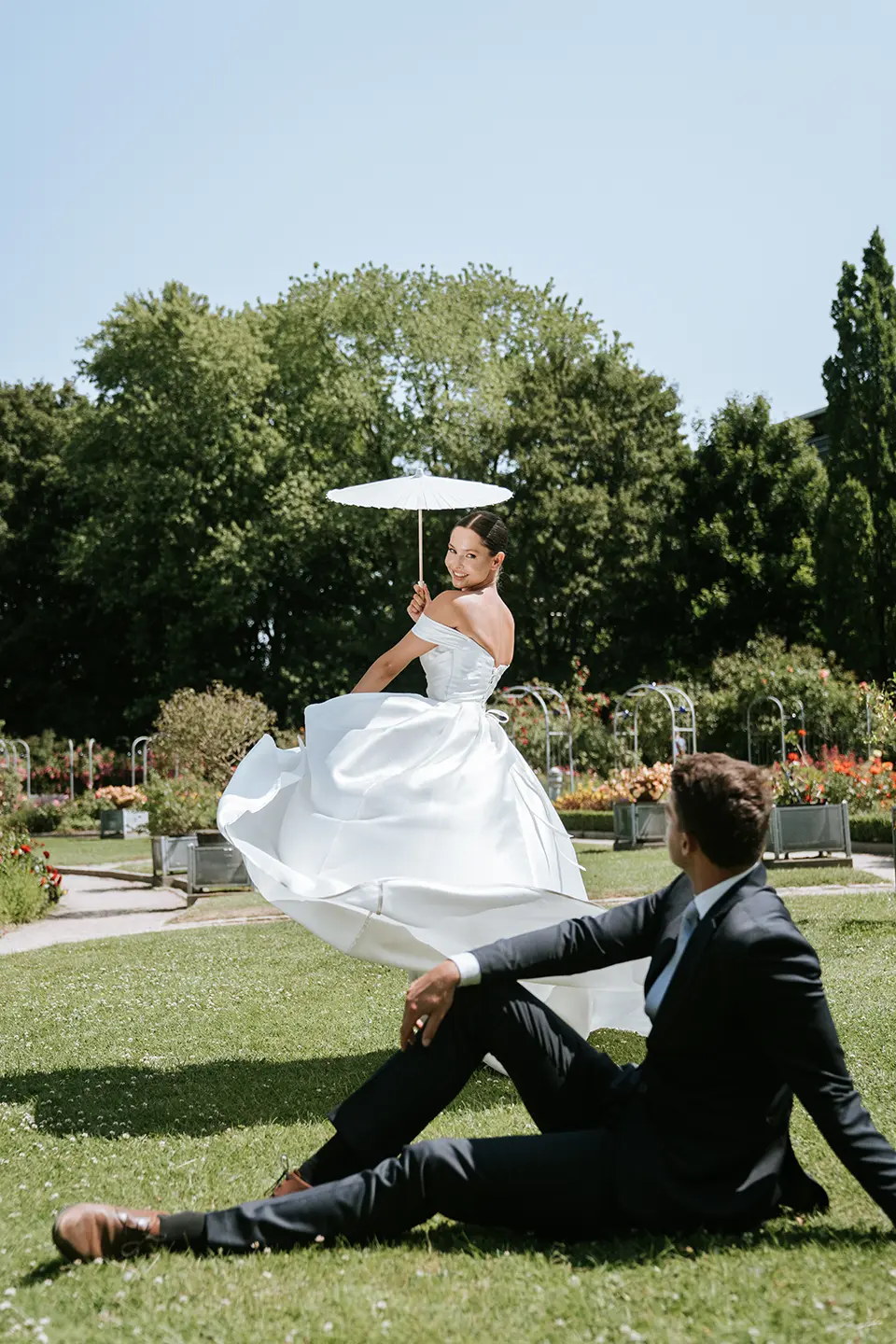 Auf der Wiese hebt die Braut schwungvoll das Kleid und hält den Parasol, vorn sitzt der Bräutigam; kreative hochzeitsfotografie und hochzeitsfotograf für zwei im Grünen