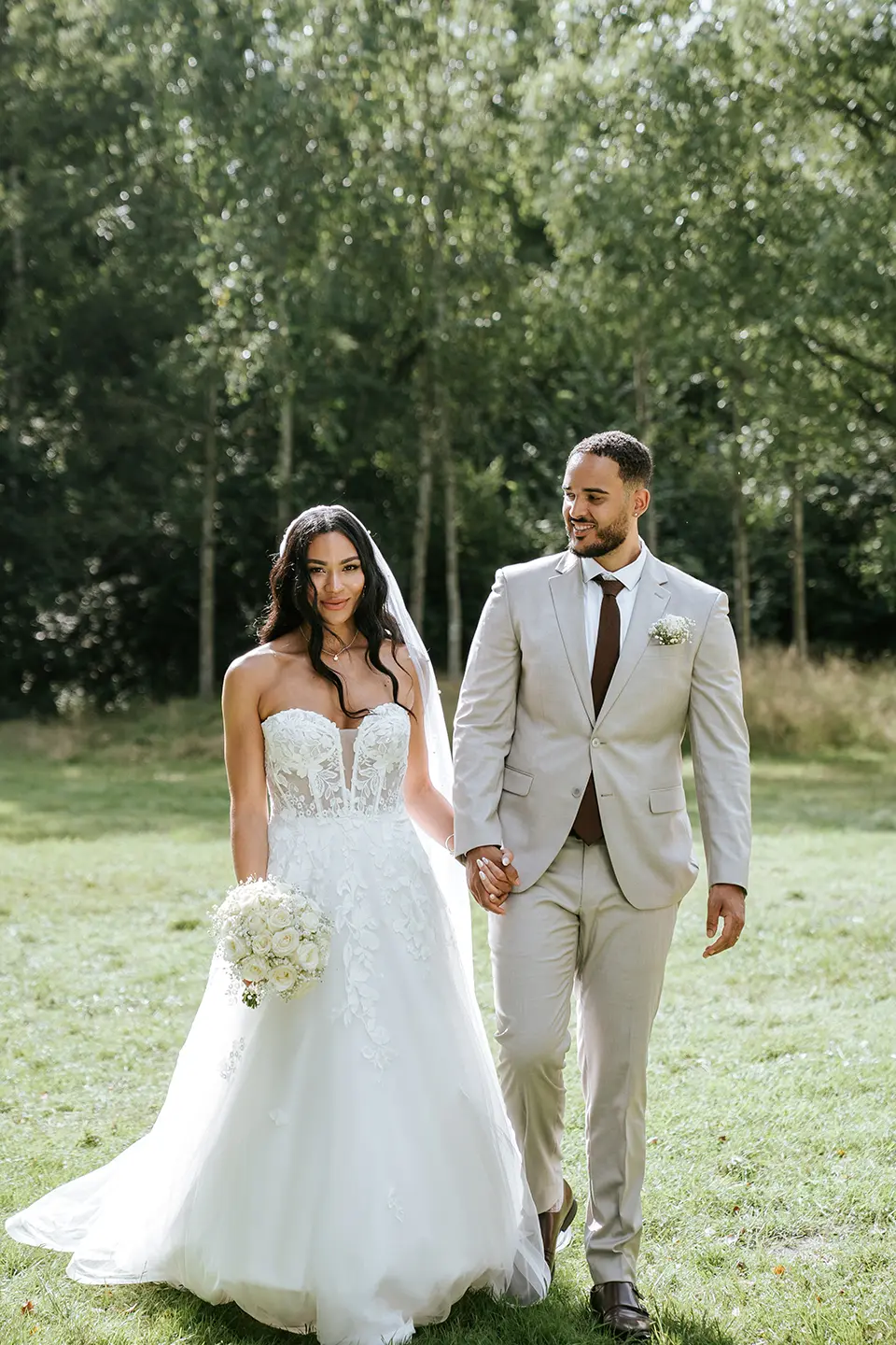 Das Paar spaziert über eine sonnige Wiese, sie im Spitzenkleid mit weißem Strauß, er im hellen Anzug. Natürliche Hochzeitsfotografie und hochzeitsbilder draußen mit dezentem Stil