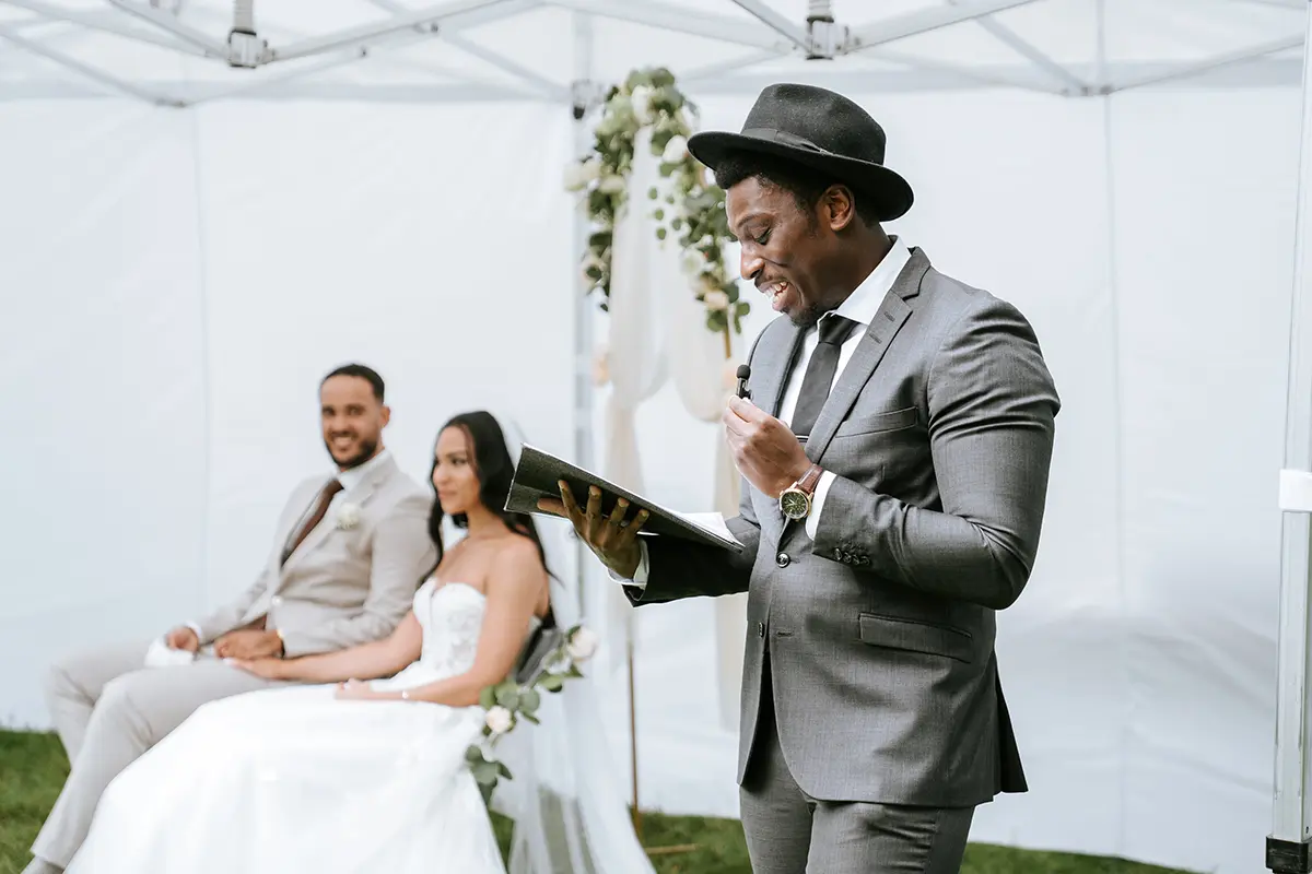 Trauredner spricht lächelnd im Zelt, das Paar sitzt dahinter Hand in Hand vor einem Blumengbogen. Reportagefotografie hochzeit und hochzeitsfotograf mit stil, ideal für emotionales hochzeitsvideo