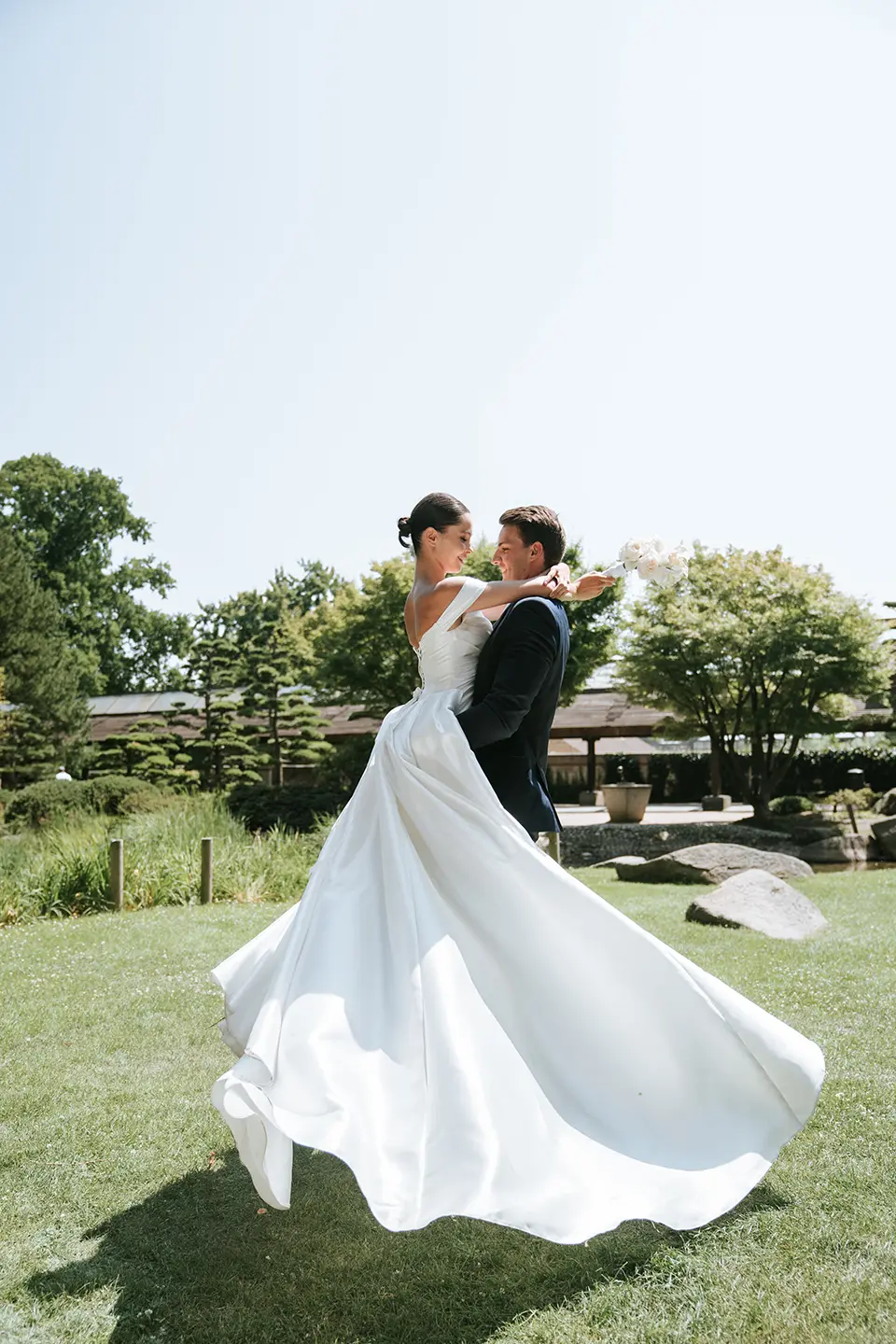 Bräutigam hebt die Braut im weiten Kleid, der Stoff wirbelt im sonnigen Park; inniger Moment in Bewegung, natürliche hochzeitsfotografie und emotionale hochzeitsbilder