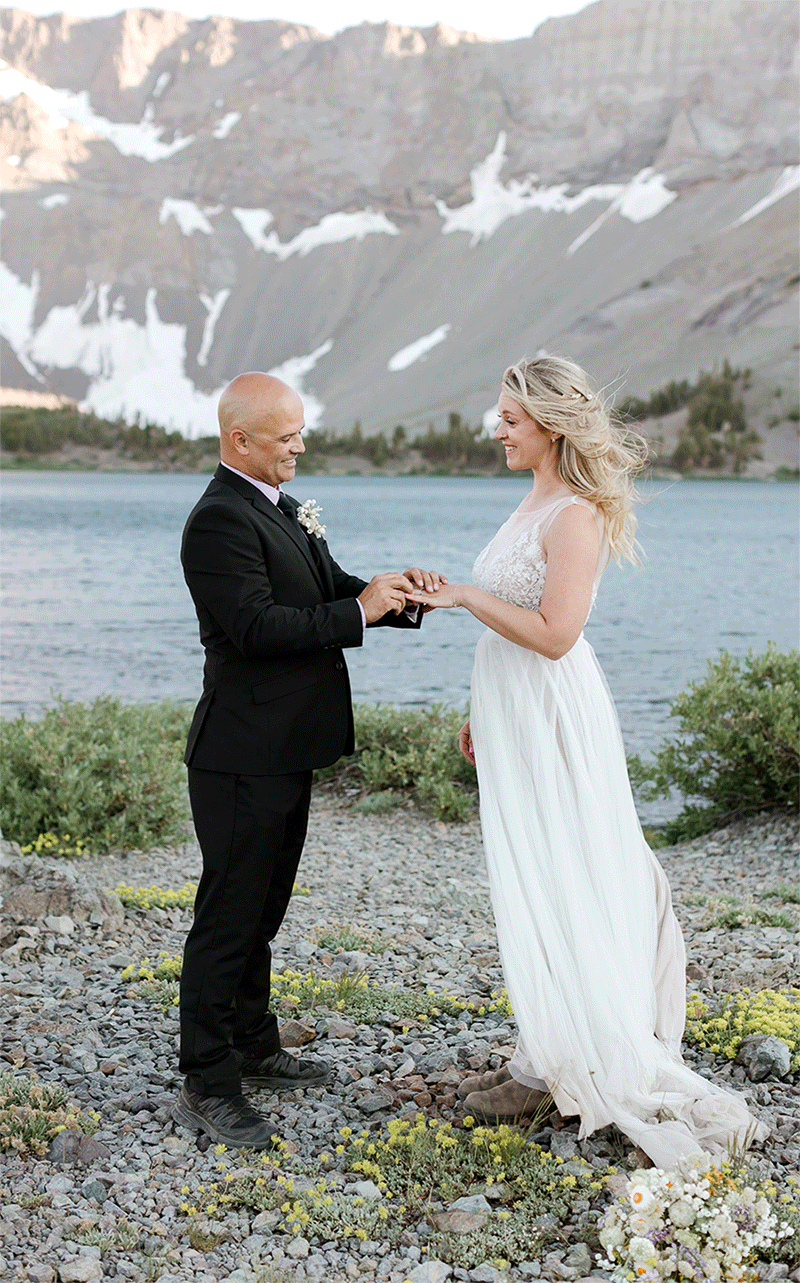 A groom slips a wedding ring on his bride's hands during their wedding ceremony on their mountain wedding day. 