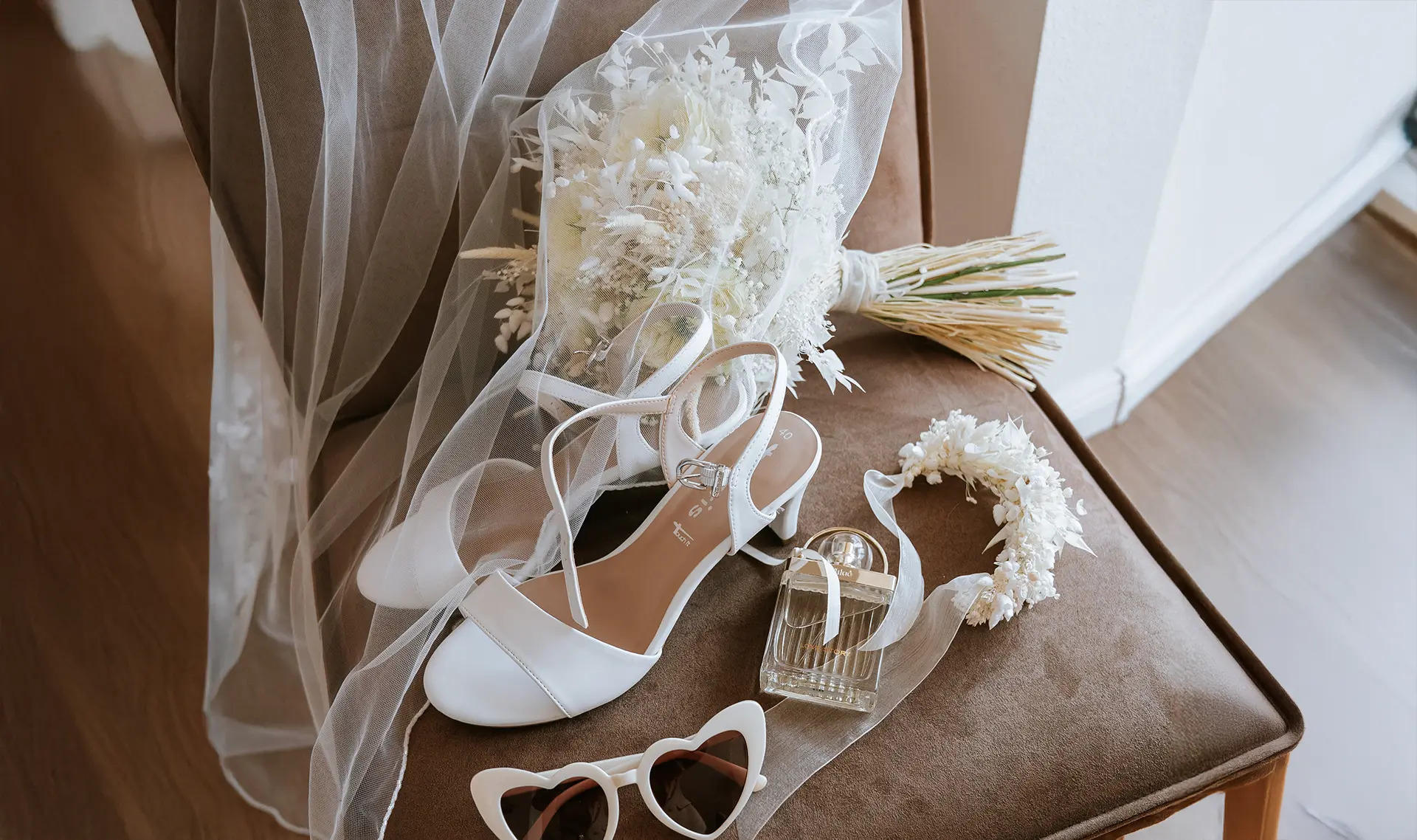 Auf dem Stuhl liegen weiße Sandalen, Schleier, getrockneter Strauß, Parfum und Herzbrille. Getting ready fotografie trifft kreative hochzeitsfotografie und setzt auf hochzeitsfotografie mit stil