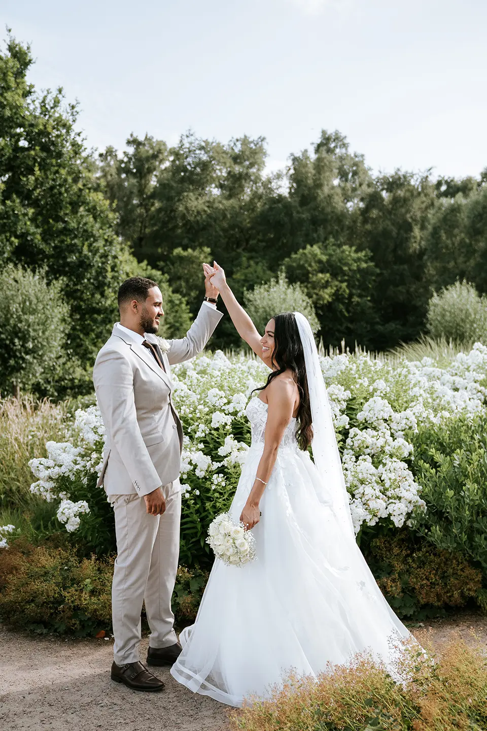 Neben üppigen Stauden hebt der Bräutigam die Hand der Braut zum Dreh. Kreative Hochzeitsfotografie trifft auf moderne Hochzeitsfotos in einer leichten, romantischen Szene
