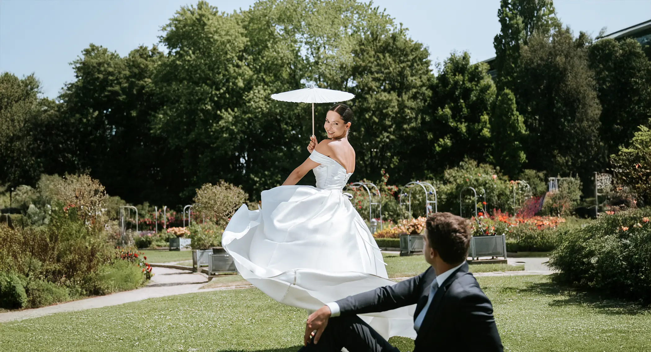 Die Braut wirbelt mit weißem Parasol durch die Parkwege, der Bräutigam schaut lächelnd zu; moderne hochzeitsfotografie und hochzeitsbilder draußen im Sommerlicht