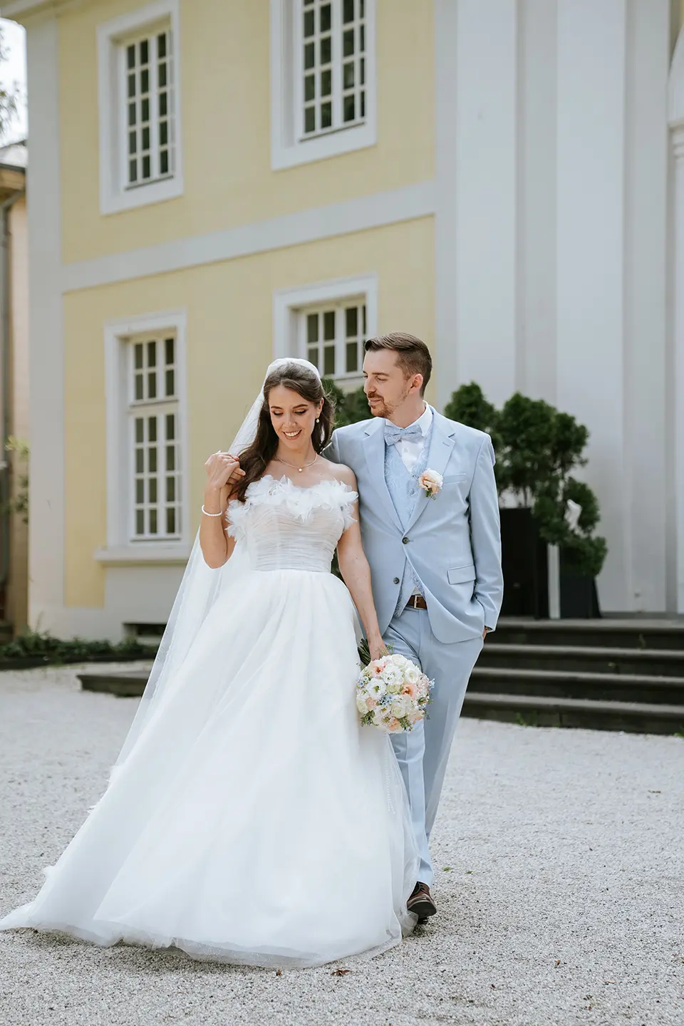 Braut und Bräutigam spazieren vor gelb-weißer Villa, lächelnd Arm in Arm mit Bouquet; reportagefotografie hochzeit und natürliche hochzeitsfotografie prägen den Moment