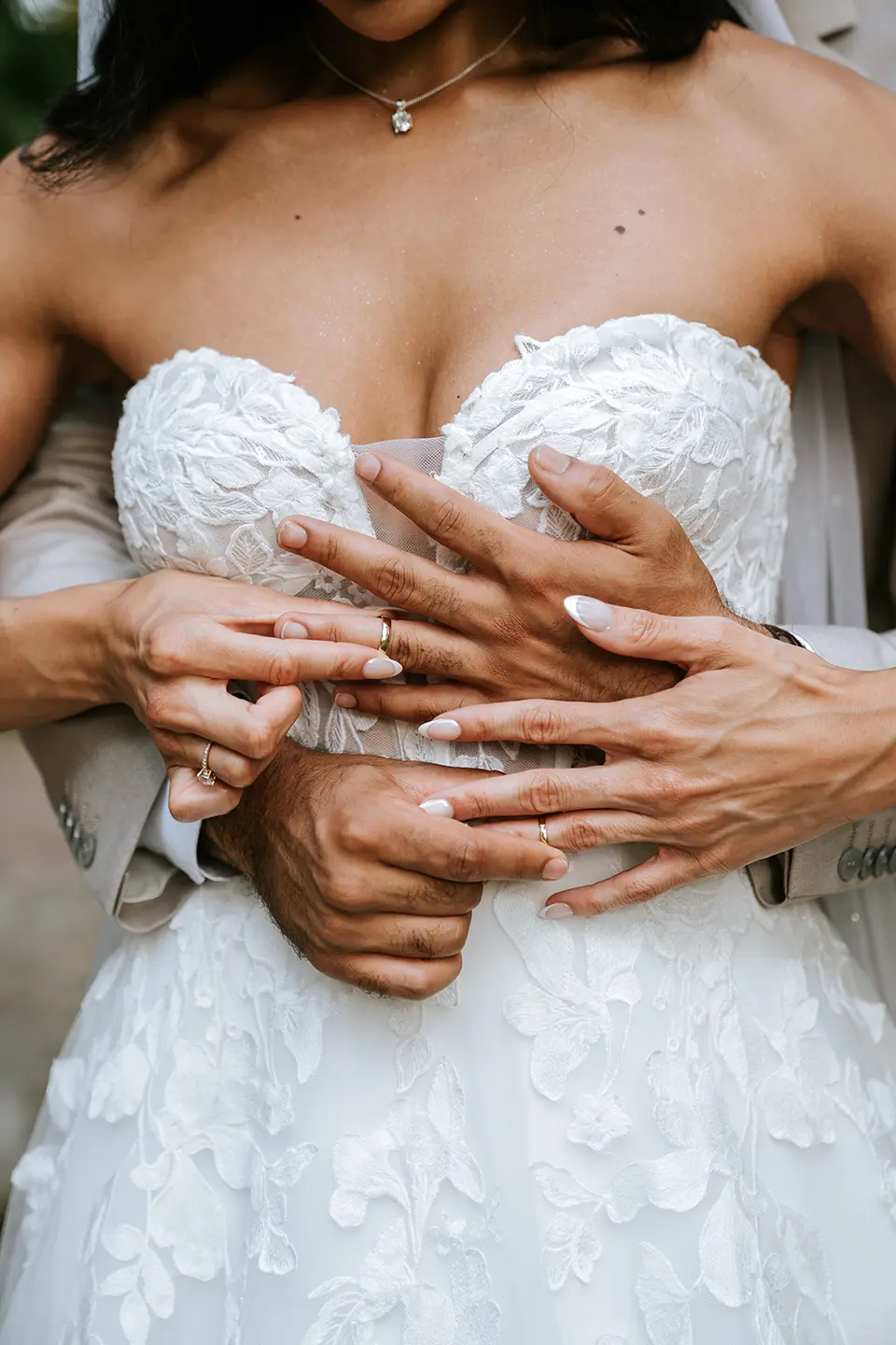 Nahaufnahme von ineinandergelegten Händen über Spitze, Eheringe glänzen vor dem Korsagenkleid. Künstlerische Hochzeitsfotos und moderne Hochzeitsfotografie im Fokus