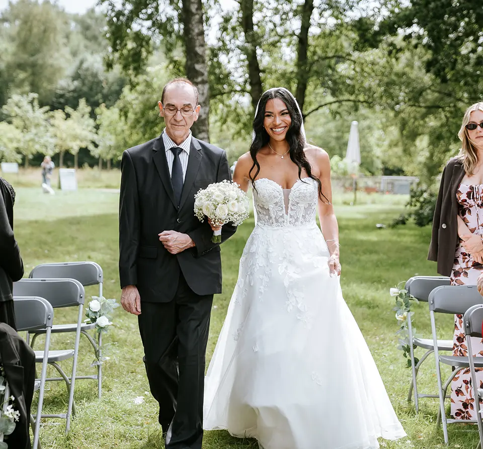 Braut schreitet mit Begleiter den Mittelgang entlang, Gäste stehen auf und lächeln, Bäume spenden Schatten. Reportagefotografie hochzeit hält emotionale hochzeitsbilder authentisch fest