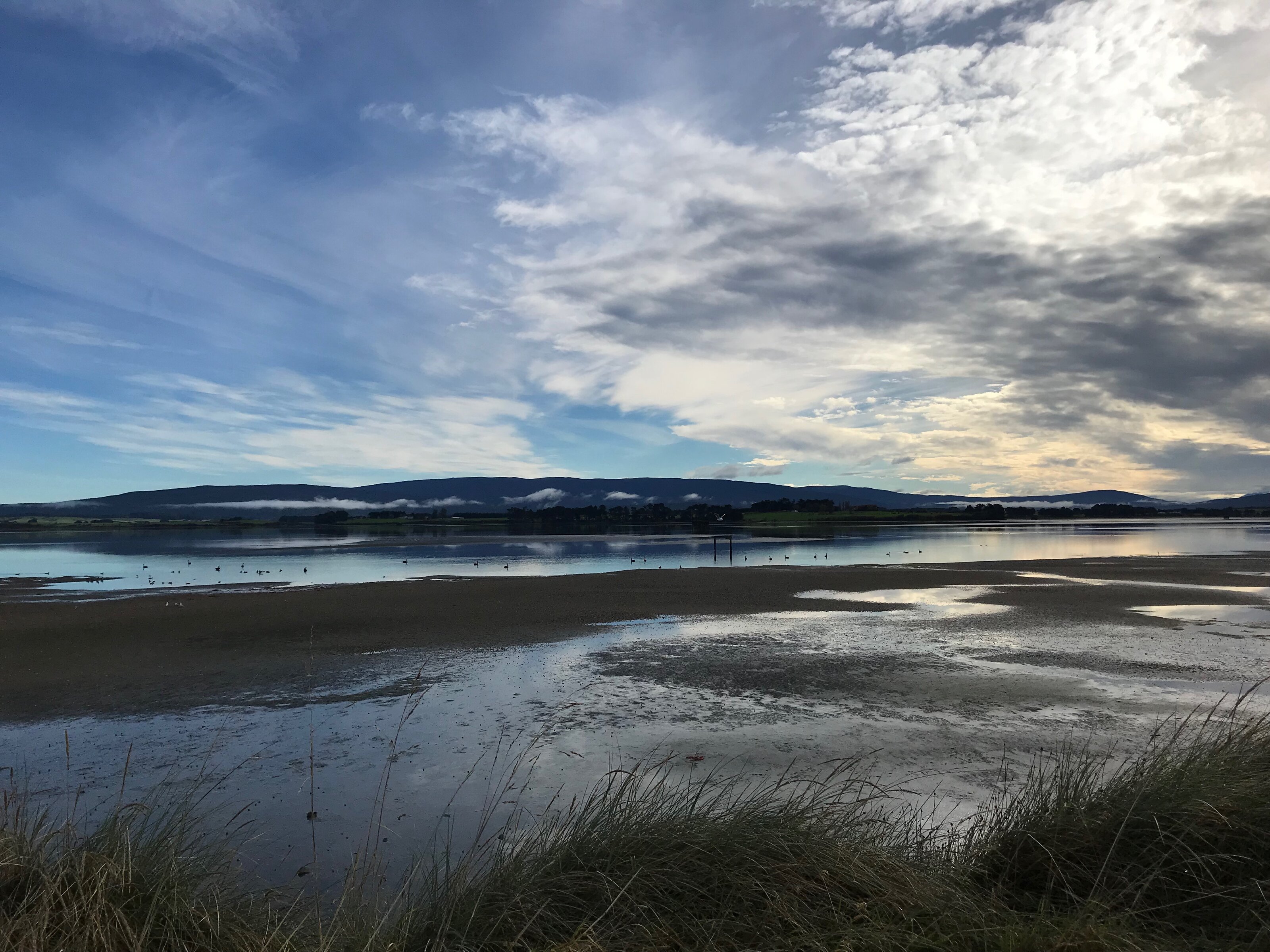 View across the Pleasure Bay Lagoon on the Estuary Walkway in Invercargill, with still water, sand flats, and low hills in the distance.