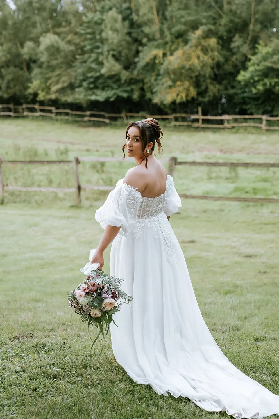 Braut in Spitzenkleid blickt über die Schulter, mit Bouquet und langer Schleppe auf der Wiese – natürliche Hochzeitsfotografie und Hochzeitsbilder draußen vom hochwertiger Hochzeitsfotograf Kiel