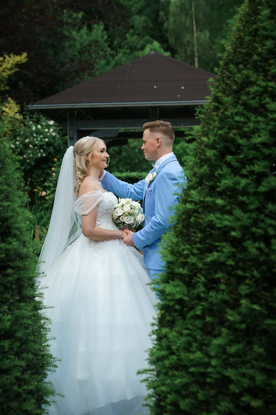 Braut und Bräutigam stehen sich liebevoll im Garten gegenüber; weißes Kleid und blauer Anzug inmitten grüner Bäume – romantische hochzeitsreportage lindern mit natürlicher hochzeitsfotografie