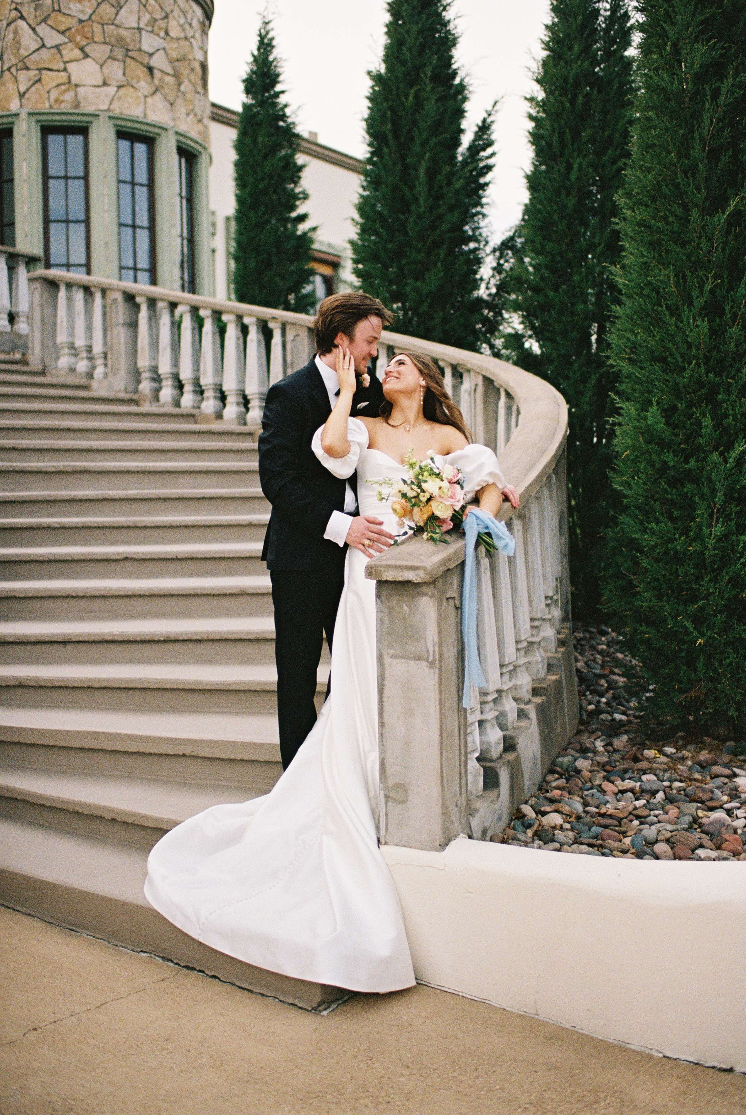 A man and woman are posing together on a set of stairs. The woman is dressed in an elegant wedding gown, complete with a bridal veil. The scene is set outdoors, creating a beautiful backdrop for this special moment.