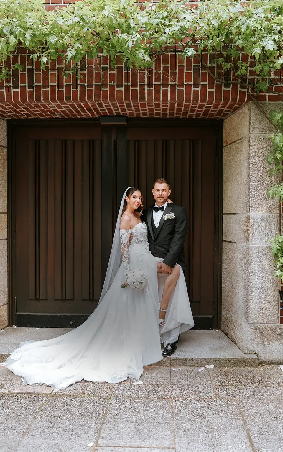 Vor dem dunklen Portal hebt der Bräutigam seine Braut mit Schleppe; Efeu und Backstein rahmen die Szene, natürliche hochzeitsfotografie und hochzeitsfotograf für zwei