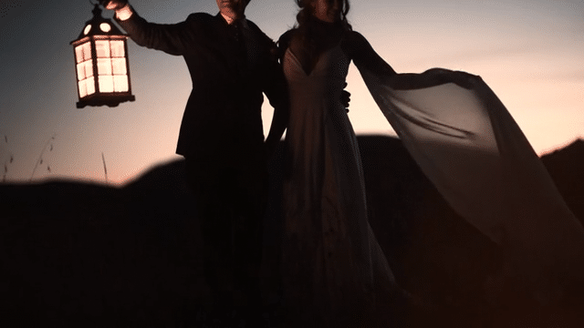 A bride and groom stand in a side hug with the groom holding a lit lantern. There is light behind them, but the sun has set and there is only a glow behind the hill. The bride is holding her train as it blows in the wind